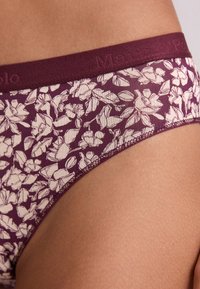 Gros plan sur un sous-vêtement floral violet sur la peau, avec une ceinture texturée et un logo discret.
