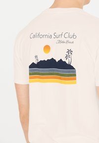 Vit bomullströja med ett tryckt mönster av berg, palmer, en sol och färgglada horisontella ränder i blått, grönt, orange och gult.