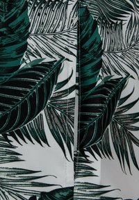Tissu à motif présentant de grandes feuilles tropicales vertes et noires sur fond blanc, avec un détail de couture bien visible le long de la couture.