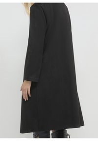 Femme portant un long manteau noir à manches droites et des bottes noires mi-mollet, debout devant un fond clair uni.