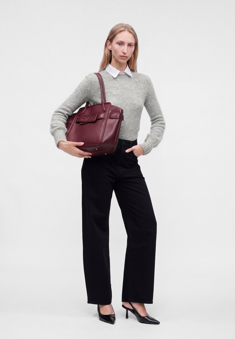 Jeune femme en pull gris, chemise blanche à col, pantalon noir et talons tenant un grand sac à main en cuir bordeaux sur fond blanc.