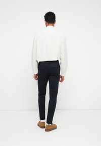 Cămașă albă cu dungi, cu nasturi, purtată cu pantaloni bleumarin și pantofi bej. Cămașa are mâneci lungi și guler standard.