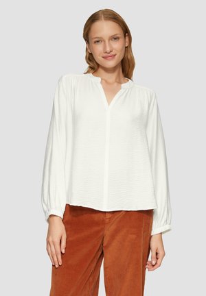 Blouse blanche à manches longues avec un col à patte, épaules froncées et poignets élastiques. Associée à un pantalon en velours côtelé de couleur rouille.