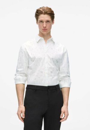 Jeune homme aux cheveux tirés en arrière, portant une chemise blanche à manches longues boutonnée et un pantalon noir, debout devant un fond blanc uni.