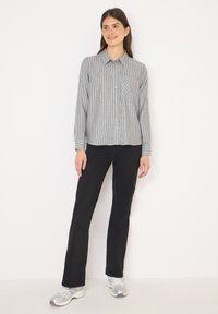 Vaaleanharmaa raidallinen nappipaita, jossa on tasku, yhdistetty mustiin farkkuihin ja hopeisiin urheilukenkiin, seisoo yksinkertaista taustaa vasten.