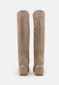 Bottes en daim beige jusqu'au genou avec une texture lisse, fermeture éclair et talon carré agrémenté d'accents métalliques. Vue de dos.