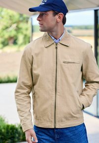Beige lightweight jacket with a zip front, pointed collar, and side pockets; worn over a blue shirt. Dark blue cap featured.