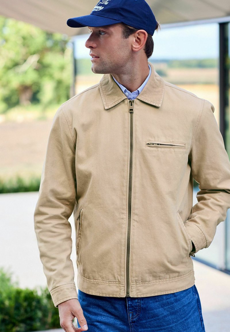 Beige lightweight jacket with a zip front, pointed collar, and side pockets; worn over a blue shirt. Dark blue cap featured.