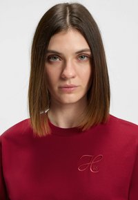 Rød bomuldssweatshirt med rund halsudskæring og et tekstureret broderet logo i en mørkere nuance. Glat, medium-vægtigt stof.