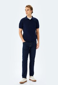 Marinblå kortärmad polo med ribbad textur, knappad krage, i kombination med mörka jeans och beige sneakers. Enkelt, avslappnat design.