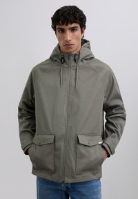 Chaqueta impermeable verde oliva con capucha, cierre de cremallera y dos bolsillos frontales. Presenta cordones ajustables y una textura suave.