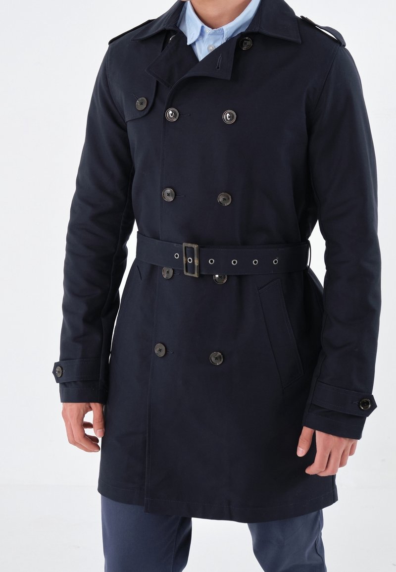 Homme portant un trench-coat bleu marine à double boutonnage avec ceinture, poignets boutonnés et poches avant, par-dessus une chemise bleu clair à col.