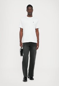 Hvit bomulls t-skjorte med en liten logo, kombinert med mørkegrå rette jeans og svarte sko, holder en svart veske. Enkelt design.
