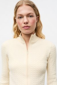 Kremfarget genser med glidelås foran, med teksturert kabelstrikkmønster, ribbet krage og lange ermer.