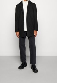 Černé strukturované sako přes bílý rolák, doplněné tmavými džínovými kalhotami a černými masivními botami. Má dva knoflíky a klopu s výřezem.