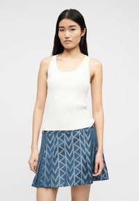 Νεαρή γυναίκα με μακριά μαύρα μαλλιά που φοράει λευκό χωρίς μανίκια ριμπ τοπ και μπλε πλισέ φούστα με γεωμετρικό μοτίβο.