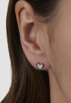 Goldene und silberne herzförmige Ohrstecker mit glattem weißem Emaille-Zentrum, gezeigt in einer Nahaufnahme eines Ohrs.