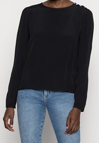 Musta pitkähihaine paita pyöreällä kaula-aukolla, jossa on koristeelliset napit olkapäillä ja sileä kangas. Yhdistetty sinisiin farkkuihin.