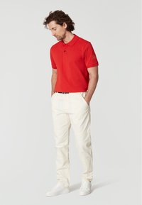 Röd polo skjorta med krage och korta ärmar, kombinerad med krämfärgade byxor och vita sneakers, stående mot en neutral bakgrund.