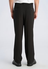 Mand iført sorte habitbukser og sorte sko, stående med ryggen mod beskueren foran en ensartet lys baggrund.