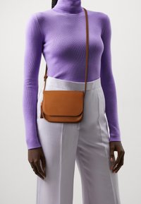 Bolso bandolera de cuero marrón con cierre de solapa; textura lisa y correa ajustable. Llevado sobre una camiseta de manga larga morada y pantalones claros.