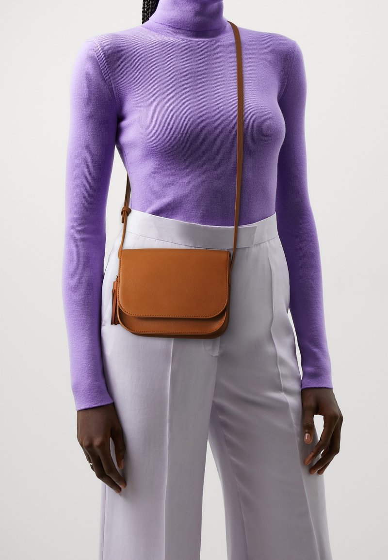 Bolso bandolera de cuero marrón con cierre de solapa; textura lisa y correa ajustable. Llevado sobre una camiseta de manga larga morada y pantalones claros.