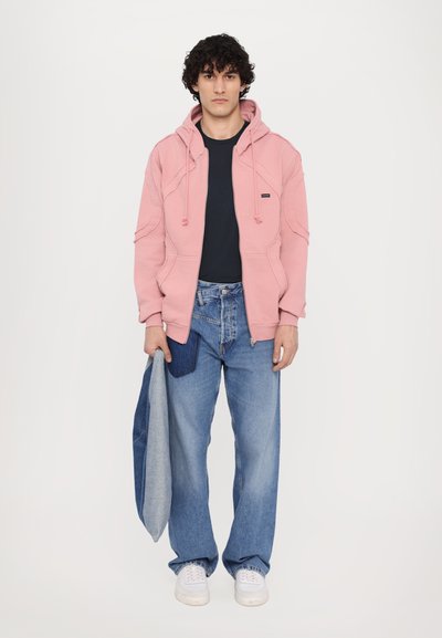 Helle rosa Kapuzenpullover mit Kordelzug, kombiniert mit einem marineblauen Shirt und verblassten, weit geschnittenen blauen Jeans. Weiße Turnschuhe und eine graue Jeansjacke.