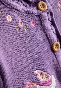 Lila gestrickte Strickjacke mit Holzknöpfen, die eine florale Stickerei in Pink und Grün auf der Vorderseite aufweist. Weiche Textur, gerippte Kanten.