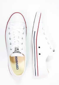 Bílé plátěné nízké tenisky s bílými tkaničkami, gumovou špičkou, červeno-námornickou pruhovanou podešví a logem "CONVERSE" na stélce.