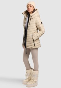 Giacca puffer beige con motivo a righe orizzontali, un ampio cappuccio e zip nere. Abbinata a leggings beige chiaro e stivali da neve crema.