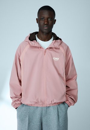 Mladi moški, oblečen v svetlo roza jopico z zadrgo in kapuco, nad belo majico in sivimi hlačami, stoji pred enobarvnim ozadjem.