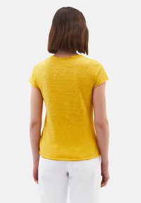 Haut jaune à manches courtes avec un col rond et un ourlet légèrement courbé. Le tissu semble doux avec une texture subtile. Pantalon blanc visible.