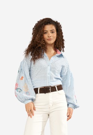 Noor naine lokkis juustega, kandes helehalli ruudulist pluusi, millel on tikitud mereloomad puffsärkidel, ning valgeid pükse pruuni vööga.