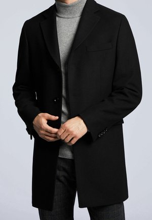 Homme portant un manteau noir simple boutonnage sur un pull à col roulé gris, ajustant les boutons du manteau, sur un fond clair uni.
