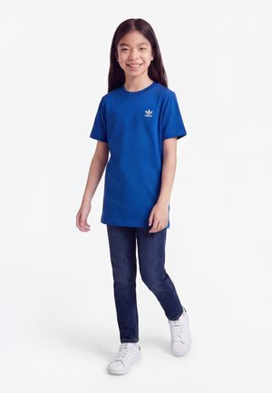 Lány sétál és mosolyog, kék Adidas pólót, sötét farmert és fehér sportcipőt visel, egyszínű fehér háttér előtt.