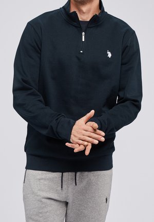 Homme portant un sweat-shirt noir à fermeture éclair quart, pantalon de survêtement gris, mains jointes devant, sur un fond clair uni.