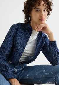 Camicia blu navy con complessi motivi floreali azzurri, abbinata a un top metallico argento e jeans azzurri chiari decorati con borchie argentate.