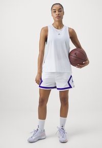 Valge ilma varrukateta sportlik topp musta logoga, koos valgete lühikestega, millel on lilla äär ja aksessorid, valged sokid ja mitmevärvilised tossud.
