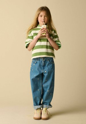 Enfant aux longs cheveux portant un t-shirt rayé vert et blanc et un jean bleu avec des motifs floraux, buvant dans une boîte de jus avec une paille.