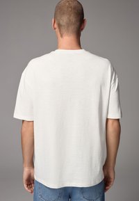Loose-fitting white cotton t-shirt with short sleeves, a crew neck, and a smooth texture, shown from the back against a gray background.
