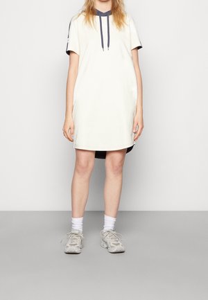 Helles, kurzärmliges Kleid mit Kapuze, das dunkelblaue Akzente an den Ärmeln und am Ausschnitt aufweist. Das Model trägt graue Sneakers und weiße Socken dazu.