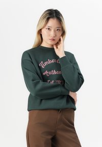 Tmavohnedá mikina s ružovým potlačeným textom "Timberland Authentic Est 1973." Má okrúhly výstrih a voľný strih. Skombinovaná s hnedými nohavicami.