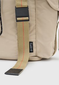 Beige nylon tas met een brede, gestructureerde schouderband met een rode accent. Zwarte plastic gesp, leren basisdetail en zichtbaar merklabel.