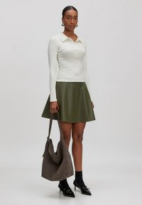 Polo shirt blanc à manches longues avec boutons, associé à une jupe évasée en faux cuir verte. Les accessoires comprennent un sac à main en daim gris et des talons noirs.