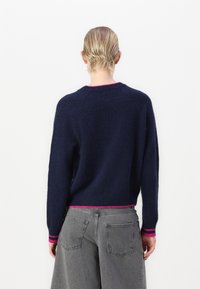 Pulover bleumarin cu un finisaj texturat, cu manșete și tiv roz cu nervuri. Purtați cu pantaloni lejeri de culoare gri deschis. Vedere din spate.