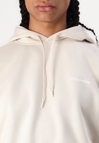 Hooded sweatshirt in light beige cotton, featuring a white “SALOMON” logo on the front. Drawstrings and soft texture evident.