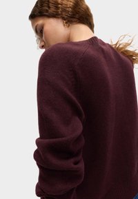 Burgundy stickad tröja med rund hals, något överdimensionerade ärmar och en texturerad tyg. Baksidan visar en ribbad halsringning och mjukhet.