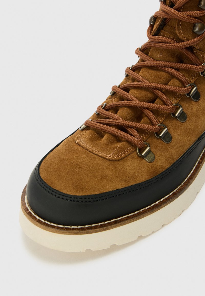 Bruine suède wandelschoen met zwart leren accenten, bruine veters, metalen veterogen en een dikke witte zool. Heeft een gestikte rand voor duurzaamheid.