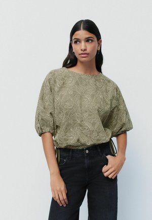 Blusa de color verde oliva con un patrón abstracto texturizado; mangas globo; puños elásticos; combinada con jeans oscuros.