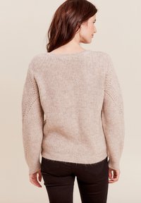 Hellbeige gestrickter Pullover mit lockerer Passform, der über eine gerippte Struktur und seitliche Details verfügt, kombiniert mit dunklen, eng anliegenden Jeans.